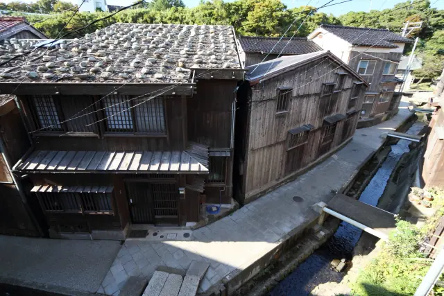 細い水路と道のそばに立つ板張りの家、石を置いた屋根